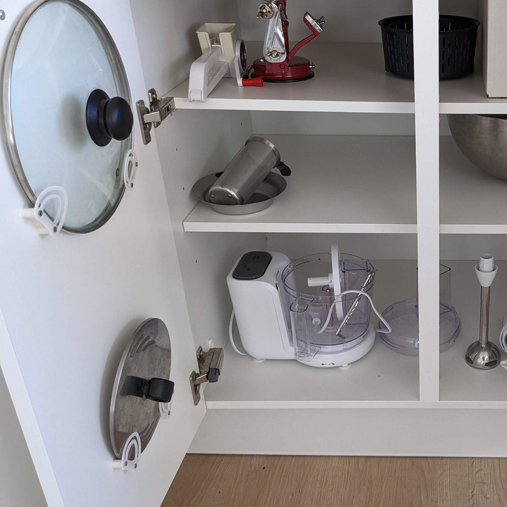 Pot lid holder clips mounted inside a kitchen cabinet door holding a glass lid, view 2