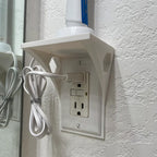 Outlet shelf mounted over a wall outlet holding a phone while it charges, view 2